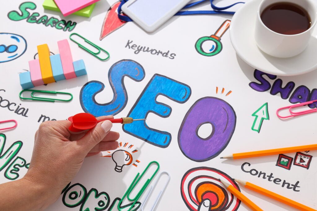 SEO A person holds a dart over a colorful Burnesseo SEO strategy diagram, featuring keywords, social media, and content, alongside a coffee cup, pencils, and sticky notes.