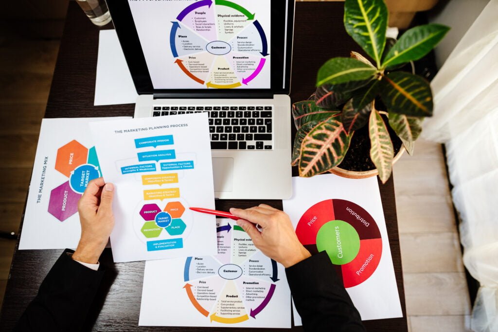 SEO A person from Burnesseo SEO Services holds a marketing planning process chart at a desk, where a laptop displays colorful diagrams. Nearby, a plant and papers with graphs complete the scene.