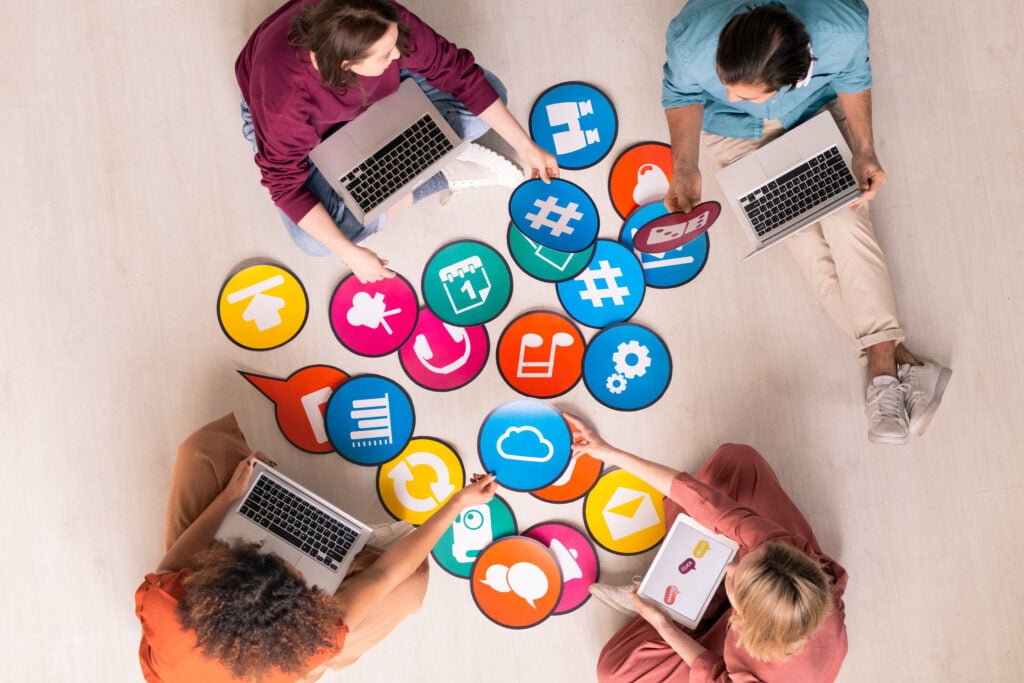 SEO Four people sit on the floor with laptops, surrounded by large icons of social media and online communication symbols, brainstorming innovative strategies. They're powered by Burnesseo SEO services to boost their online presence effectively.