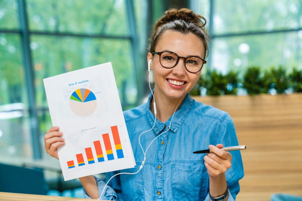 Young successful smart lady showing diagrams holding pen while using headphones in modern office