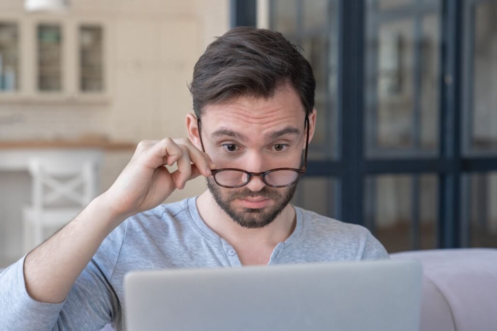 SEO A man with glasses looks at a laptop screen with a focused expression, pulling his glasses down slightly. He is indoors with a kitchen and glass doors in the background.